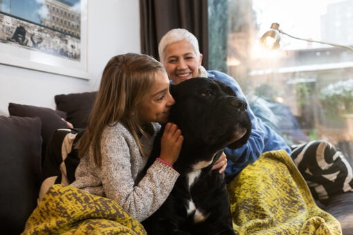 Niña abrazando a un perro negro grande sobre un sofá, con una mujer de cabello corto al fondo sonriendo